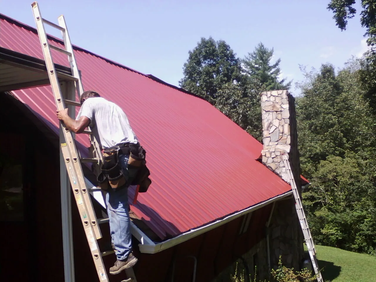 Roofer on a ladder beside a red metal roof and stone chimney — Soffit & Fascia Repair by Roofing Brothers in Mc Gill